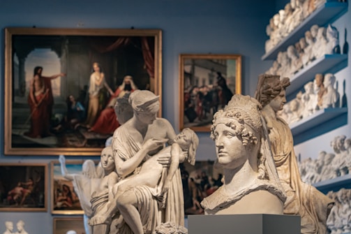 A room filled with classical sculptures and paintings. Busts and full-sized statues are intricately crafted, depicting figures in flowing robes and detailed headgear. Paintings hang on the walls, showing historical or mythological scenes with rich, deep colors.