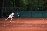 A tennis player in motion, executing a serve on a clay court. The background features a green fence and dense foliage, while the player is in athletic gear, focusing intently on the ball.