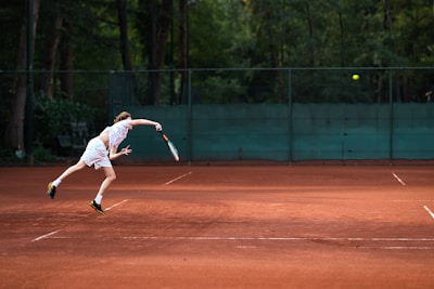 A tennis player in motion, executing a serve on a clay court. The background features a green fence and dense foliage, while the player is in athletic gear, focusing intently on the ball.