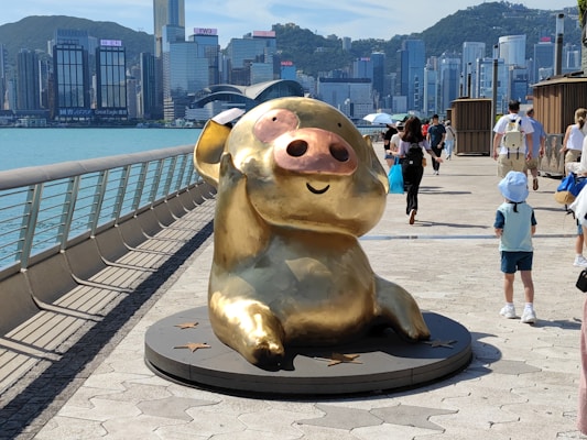 A large, shiny gold sculpture of a cartoon-like animal sits on a platform with star decorations. It is positioned on a waterfront promenade with people walking by, some with umbrellas. In the background, a city skyline featuring tall buildings and hills is visible across the water.