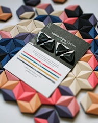 Several geometric, three-dimensional shapes are arranged in an overlapping pattern, featuring a variety of colors including black, blue, pink, and beige. In the center, there is a card labeled 'ROMBO PRISMA ECO BLACK' showcasing three black triangular objects with white accents. The card also includes some descriptive text and a colorful graph at the bottom.