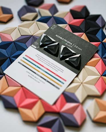 Several geometric, three-dimensional shapes are arranged in an overlapping pattern, featuring a variety of colors including black, blue, pink, and beige. In the center, there is a card labeled 'ROMBO PRISMA ECO BLACK' showcasing three black triangular objects with white accents. The card also includes some descriptive text and a colorful graph at the bottom.