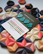A flat lay display featuring a set of colorful geometric tiles arranged in a hexagonal pattern. In the foreground, there is a card with text and graphics, accompanied by three turquoise picks that resemble guitar picks. The geometric tiles are in shades of pink, beige, blue, and black, creating a visually striking contrast with the card and picks.