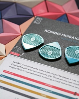 The image features two stylish guitar picks with intricate patterns, placed on a textured gray surface. A description of the guitar picks' features is visible below them, alongside a color-coded chart evaluating aspects such as release, attack, and flexibility. In the background, there is a vibrant mosaic with geometric shapes in shades of pink, blue, and beige.