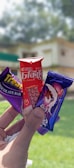 A hand holding three chocolate bars with colorful packaging. The chocolates are held against a blurred background of greenery and a building. The packages are mainly red, violet, and purple with distinct branding and illustrations of chocolate.