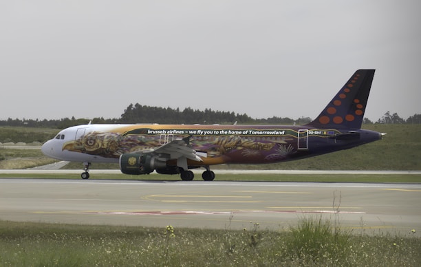 A commercial airplane with elaborate artwork on its body is taxing on a runway. The plane features colorful murals and text that reads 'we fly you to the home of Tomorrowland.' The background includes a partially cloudy sky, grassy fields, and trees in the distance.
