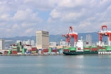 A bustling seaport with a large cargo ship labeled 'EVERGREEN' docked near the shore. Numerous colorful shipping containers are stacked near the water. The background features several high-rise buildings, mountains, and a partly cloudy sky. Large red cranes are present, used for loading and unloading containers.