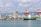 A bustling seaport with a large cargo ship labeled 'EVERGREEN' docked near the shore. Numerous colorful shipping containers are stacked near the water. The background features several high-rise buildings, mountains, and a partly cloudy sky. Large red cranes are present, used for loading and unloading containers.
