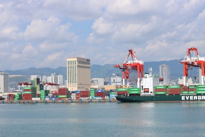 A bustling seaport with a large cargo ship labeled 'EVERGREEN' docked near the shore. Numerous colorful shipping containers are stacked near the water. The background features several high-rise buildings, mountains, and a partly cloudy sky. Large red cranes are present, used for loading and unloading containers.