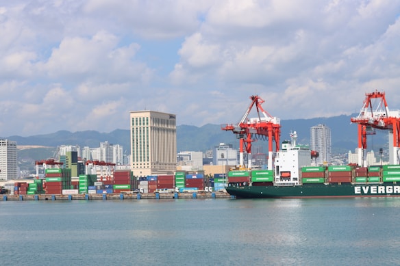 A bustling seaport with a large cargo ship labeled 'EVERGREEN' docked near the shore. Numerous colorful shipping containers are stacked near the water. The background features several high-rise buildings, mountains, and a partly cloudy sky. Large red cranes are present, used for loading and unloading containers.