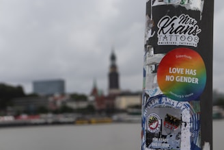 A street pole is covered with a variety of stickers, including a round rainbow sticker with the text 'LOVE HAS NO GENDER', and another sticker for a tattoo artist. The background shows an urban skyline with a prominent tower and several buildings, blurred to emphasize the pole and stickers in the foreground. The sky is overcast.