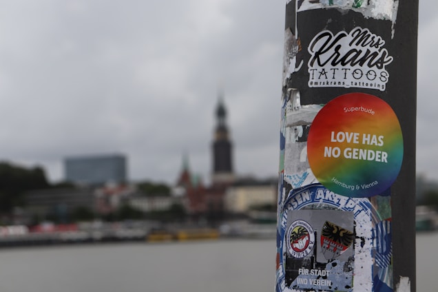 A street pole is covered with a variety of stickers, including a round rainbow sticker with the text 'LOVE HAS NO GENDER', and another sticker for a tattoo artist. The background shows an urban skyline with a prominent tower and several buildings, blurred to emphasize the pole and stickers in the foreground. The sky is overcast.