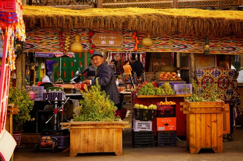 A bustling market stall adorned with a colorful fabric canopy. An elderly man in a hat adjusts items around him. Fruits like grapes, apples, and melons are neatly arranged in wooden and plastic crates. Decorative items hang from the canopy, adding a cultural touch. In the background, a green door and passing people enhance the lively atmosphere.