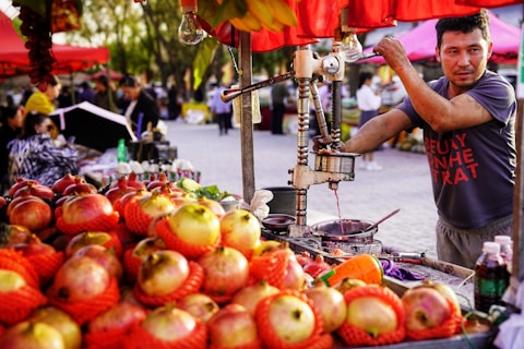 A vendor operates a juice press surrounded by neatly stacked pomegranates wrapped in red netting. The scene is vibrant with outdoor market stalls in the background, bustling with people. The vendor is focused on his task amidst a colorful and lively environment.