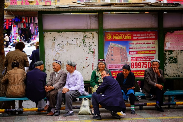 A group of people are sitting and interacting at a bus stop, wearing traditional hats and clothing. In the background, colorful clothing items are visible in a market setting, and a large poster with red text and an image of a building is displayed.
