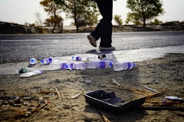 Plastic bottles are scattered across a roadside next to a person walking. The ground is sandy with some twigs and debris, while trees line the horizon. The scene appears desolate with an overcast sky.