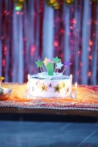 A decorated birthday cake with a colorful array of star-shaped toppers and a green candle sits on an ornate yellow and red cloth. The cake displays the name 'DEVANSH' in golden star letters, surrounded by lit candles. The backdrop features shimmering red and gold streamers.