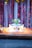 A decorated birthday cake with a colorful array of star-shaped toppers and a green candle sits on an ornate yellow and red cloth. The cake displays the name 'DEVANSH' in golden star letters, surrounded by lit candles. The backdrop features shimmering red and gold streamers.