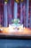 A decorated birthday cake with a colorful array of star-shaped toppers and a green candle sits on an ornate yellow and red cloth. The cake displays the name 'DEVANSH' in golden star letters, surrounded by lit candles. The backdrop features shimmering red and gold streamers.