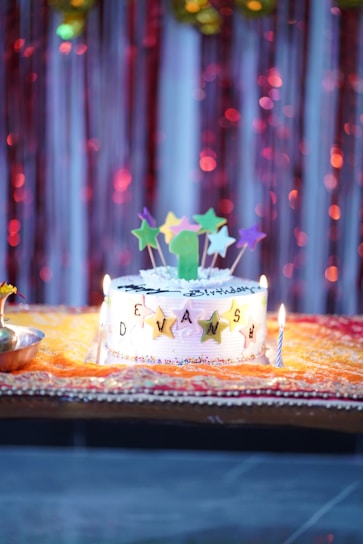 A decorated birthday cake with a colorful array of star-shaped toppers and a green candle sits on an ornate yellow and red cloth. The cake displays the name 'DEVANSH' in golden star letters, surrounded by lit candles. The backdrop features shimmering red and gold streamers.