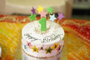 A colorful birthday cake with a number one candle and star-shaped toppers in various colors. The cake is decorated with sprinkles and has 'Happy Birthday' written on it, along with the name 'Devansh' spelled out using lettered stars. It rests on a vibrant, multicolored tablecloth that adds to the festive atmosphere.