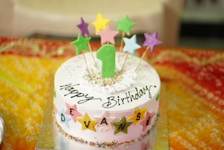 A colorful birthday cake with a number one candle and star-shaped toppers in various colors. The cake is decorated with sprinkles and has 'Happy Birthday' written on it, along with the name 'Devansh' spelled out using lettered stars. It rests on a vibrant, multicolored tablecloth that adds to the festive atmosphere.