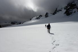 A group of climbers traverse a vast snowy landscape, leaving a trail of footprints behind. They move toward jagged mountain peaks that are partially obscured by dense, dramatic clouds.