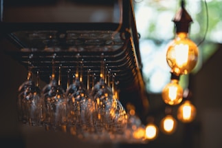 Multiple wine glasses hang upside down in a darkened bar setting, illuminated by warm, glowing light bulbs which create a soft and inviting ambiance. The foreground focuses on the glasses, with a pleasing bokeh effect in the background.