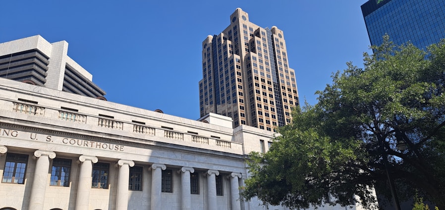A classical courthouse building with columns is in the foreground, with modern high-rise buildings rising behind it under a clear blue sky. A large, leafy tree is present on the right side of the image, adding a natural element to the urban setting.