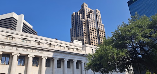A classical courthouse building with columns is in the foreground, with modern high-rise buildings rising behind it under a clear blue sky. A large, leafy tree is present on the right side of the image, adding a natural element to the urban setting.