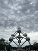 A massive structure composed of interconnected metallic spheres forming the shape of an atom stands prominently under a cloudy sky. Trees surround the monument, and a few people are visible in the foreground.