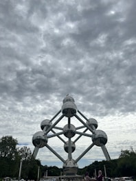 A massive structure composed of interconnected metallic spheres forming the shape of an atom stands prominently under a cloudy sky. Trees surround the monument, and a few people are visible in the foreground.