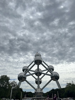 A massive structure composed of interconnected metallic spheres forming the shape of an atom stands prominently under a cloudy sky. Trees surround the monument, and a few people are visible in the foreground.