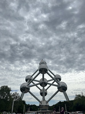 A massive structure composed of interconnected metallic spheres forming the shape of an atom stands prominently under a cloudy sky. Trees surround the monument, and a few people are visible in the foreground.
