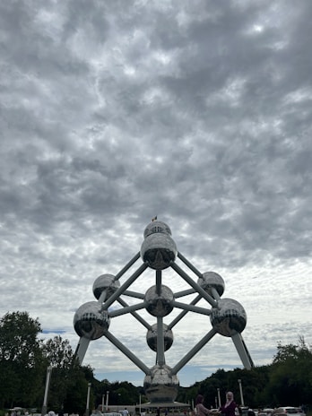 A massive structure composed of interconnected metallic spheres forming the shape of an atom stands prominently under a cloudy sky. Trees surround the monument, and a few people are visible in the foreground.