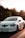 A white car is moving along a road with trees in the background and the sun setting, casting a warm glow over the scene. The car has a floral design on its side and a European-style license plate.