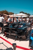 A group of people gather around a luxury sports car, possibly a Lamborghini, at an outdoor event. Numerous attendees, dressed in casual summer attire, are visible under large white umbrellas. The backdrop features green trees under a clear blue sky.