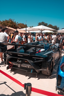 A group of people gather around a luxury sports car, possibly a Lamborghini, at an outdoor event. Numerous attendees, dressed in casual summer attire, are visible under large white umbrellas. The backdrop features green trees under a clear blue sky.