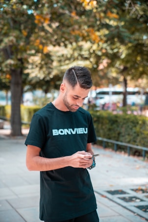 A young man with short hair wearing a black t-shirt with the word 'BONVIEU' written on it is looking down at his phone. He is standing on a paved walkway outdoors with green trees and bushes in the background.
