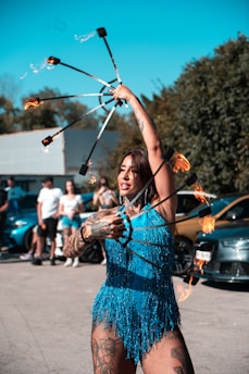 A performer is engaging an audience with a fire act, holding flaming props in each hand. The performer is wearing a vibrant blue fringed outfit and has noticeable tattoos on their arms and legs. The background features several people watching the performance and some parked cars under a clear blue sky.