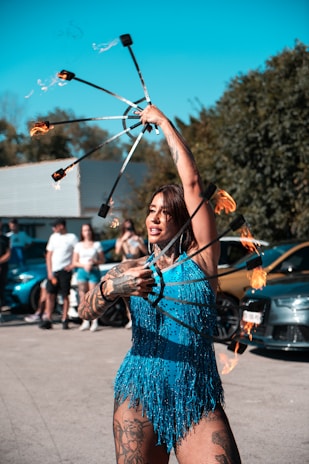 A performer is engaging an audience with a fire act, holding flaming props in each hand. The performer is wearing a vibrant blue fringed outfit and has noticeable tattoos on their arms and legs. The background features several people watching the performance and some parked cars under a clear blue sky.