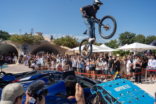 A cyclist is performing an airborne stunt over luxury sports cars in front of an audience. The crowd is gathered outdoors on a sunny day with trees and structures in the background. Many people are taking photos and videos, and there are blue skies overhead.