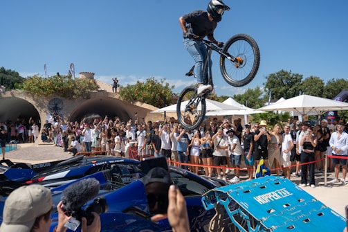 A cyclist is performing an airborne stunt over luxury sports cars in front of an audience. The crowd is gathered outdoors on a sunny day with trees and structures in the background. Many people are taking photos and videos, and there are blue skies overhead.