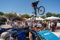 A cyclist is performing an airborne stunt over luxury sports cars in front of an audience. The crowd is gathered outdoors on a sunny day with trees and structures in the background. Many people are taking photos and videos, and there are blue skies overhead.