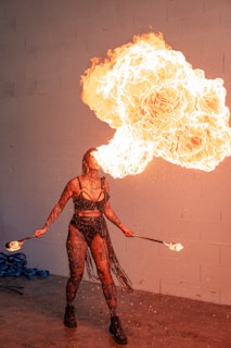 A performer is exhaling a large, dramatic burst of fire, creating a striking visual effect. The individual is dressed in a black, fringed outfit and is holding lit torches in both hands. The background is a plain wall, accentuating the intensity of the flames.