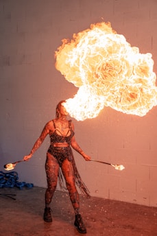 A performer is exhaling a large, dramatic burst of fire, creating a striking visual effect. The individual is dressed in a black, fringed outfit and is holding lit torches in both hands. The background is a plain wall, accentuating the intensity of the flames.