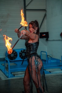 A woman dressed in a leather outfit holds two flaming batons indoors. The scene is set in an industrial space with blue machinery in the background. She has tattoos on her arms and legs and wears hoop earrings. Her expression is focused and intense.