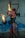 A woman dressed in a leather outfit holds two flaming batons indoors. The scene is set in an industrial space with blue machinery in the background. She has tattoos on her arms and legs and wears hoop earrings. Her expression is focused and intense.
