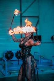 A performer is engaged in a fire dance, holding two flaming sticks with one in each hand. The individual is dressed in a studded black corset and fishnet fabric with tattooed skin visible. The performance takes place in an industrial setting with large fans in the background, and the dominant color tone is a cool blue contrasted by the fire's warm orange flames.