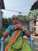 An elderly woman with grey hair sits in a blue plastic chair. She is wearing a vibrant traditional sari with a green base color and yellow floral patterns, accented with a red border. Her arms are crossed, and she is adorned with multiple bangles. The background features an outdoor setting with trees and some buildings.